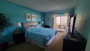 Un dormitorio con cama, mesitas de noche, escritorio, silla, planta y puerta corrediza a un balcón.