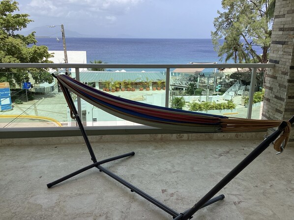 A hammock on a balcony overlooking a beach with a view of the ocean and a resort area. - Beachfront Jr Penthouse *Amazing Views*1 min walk to Beach/Restaurants Elevator (Sosúa)