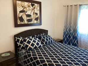 A bedroom with a patterned bedspread, a nightstand, a framed artwork on the wall, and a window with curtains. - Beachfront Jr Penthouse *Amazing Views*1 min walk to Beach/Restaurants Elevator (Sosúa)