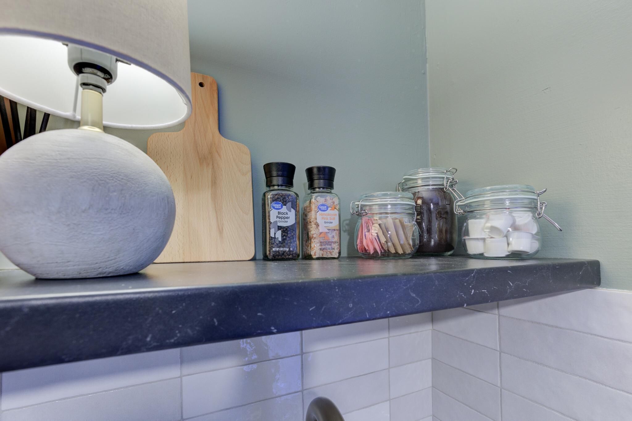 Un comptoir de cuisine avec une lampe blanche, une planche à découper en bois et divers pots à épices.