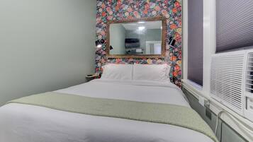 Una cama bien hecha con sábanas blancas y un plaid verde, un espejo grande encima y un estampado floral en la pared.