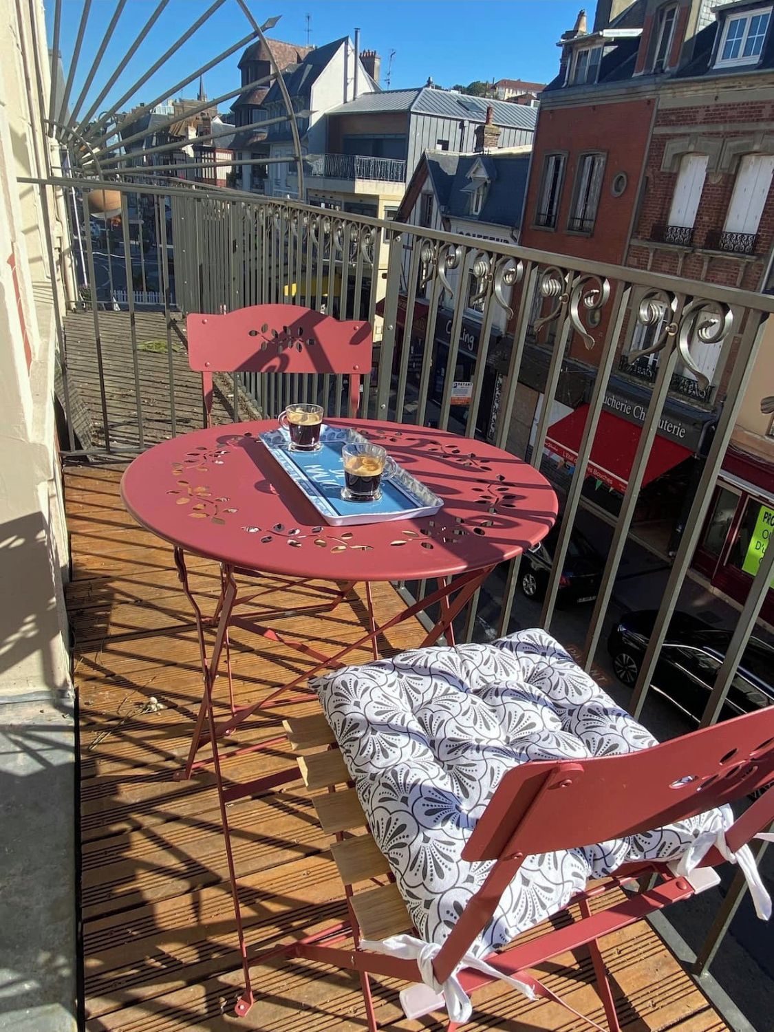 Ein roter Metalltisch mit Stühlen auf einer Holzdielenveranda, im Hintergrund sind Gebäude zu sehen.