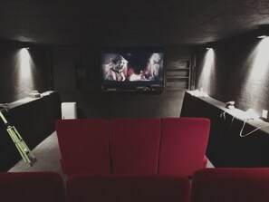 Ett mörkt rum med en platt-TV som hänger på väggen, röda sittplatser och ett litet bord med en lampa.
