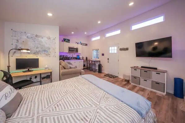 A modern hotel room with a large bed, a desk with a computer, a flat-screen TV, and a kitchenette in the background.