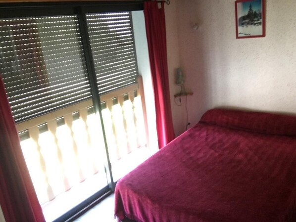 Une chambre à coucher avec un lit, une fenêtre avec des stores, un rideau rouge et un téléphone fixé au mur.