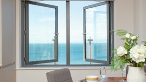 Un comedor con una mesa preparada para dos personas, un jarrón de flores y vista al mar a través de un ventanal grande.