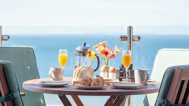 Una mesa de desayuno con vista al mar, una cafetera, dos vasos de zumo de naranja, un jarrón con flores y un croissant.