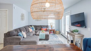 Een moderne woonkamer met een hoekbank, een houten salontafel, een grote geweven hanglamp, een flatscreen-televisie en een bijzettafeltje met een plant.