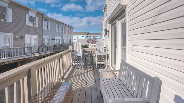 Een houten terras met witte stoelen en een tafel, met uitzicht op een woonwijk met huizen en een heldere lucht.