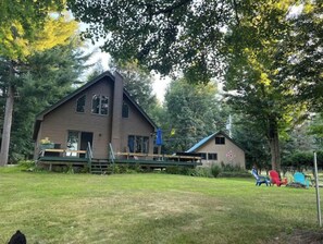 Situated in a wooded area, this cabin features a front porch with a bench and a small table, and a deck area perfect for outdoor leisure.