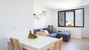A modern living room with a white dining table, wicker chairs, a grey sofa with cushions, and a large window with blinds.