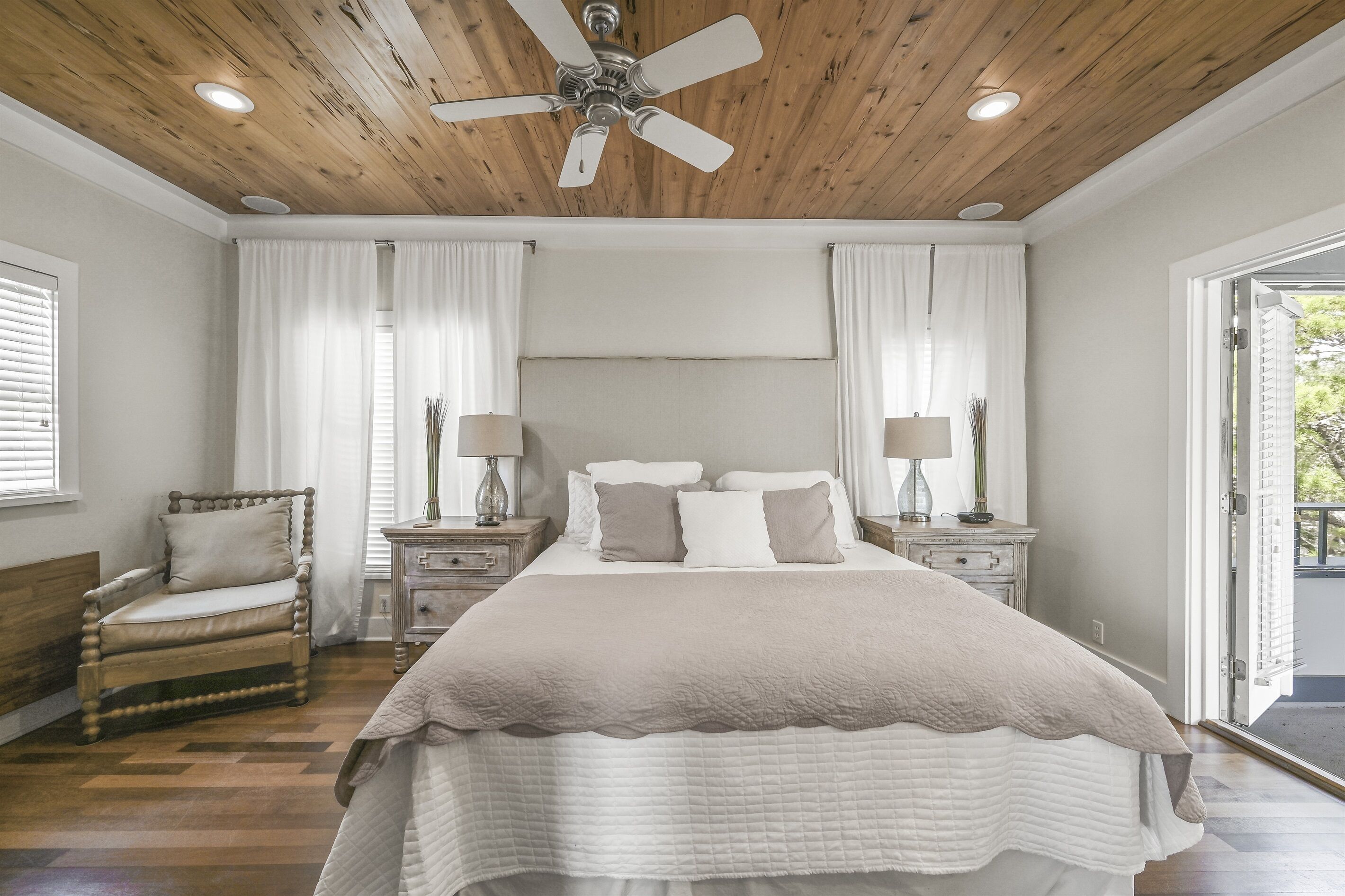 Un dormitorio con techo de madera, una cama con colcha blanca, dos mesitas de noche con lámparas, una silla y una ventana con cortinas.