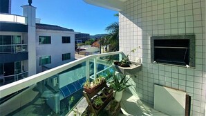 Uma varanda com guarda-corpo de vidro, plantas em vasos e vista para edifícios.
