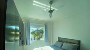 Quarto com cama, ventoinha no teto, janela com cortinas e vista para palmeiras lá fora.