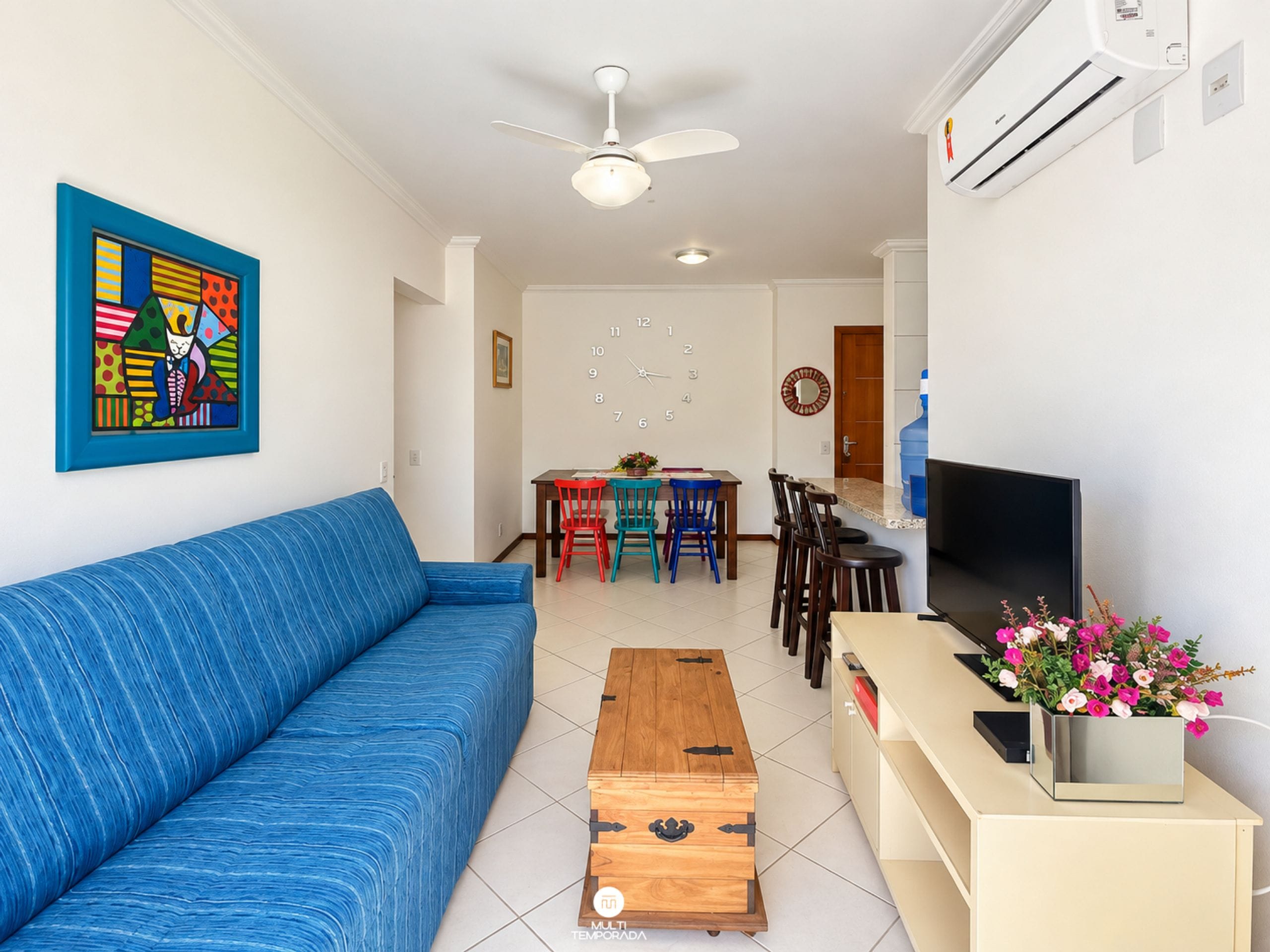 Una sala de estar con un sofá azul, una mesa de centro de madera, un comedor con sillas, un televisor y un jarrón con flores.