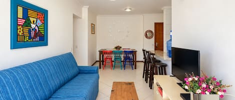 A living room with a blue sofa, a wooden coffee table, a dining area with chairs, a TV, and a flower vase.