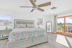 A bedroom with a bed, a ceiling fan, a nightstand, a lamp, a window with blinds, and a balcony with a view. - Private Pool, Hot Tub, Boat Dock, Putting Green (Marathon)