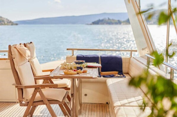Una terraza iluminada por el sol con una mesa lista para comer, asientos acolchados y vistas al mar.