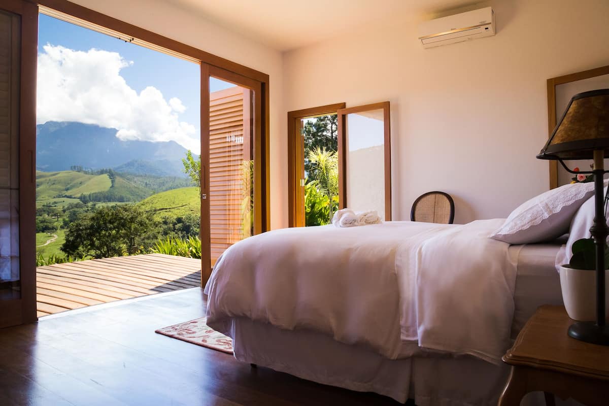 Une chambre avec un grand lit, un plancher en bois, une vue sur des collines verdoyantes et un appareil de climatisation.