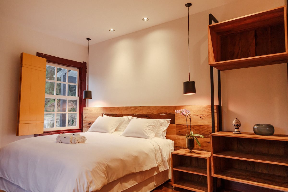 Une chambre avec une tête de lit en bois, un lit recouvert de draps blancs, une fenêtre donnant sur un beau paysage, et une table de chevet agrémentée d’un vase et d’un bol.