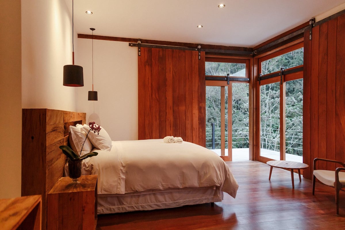 Une chambre moderne avec un grand lit, des murs en bois et une vue sur les arbres à travers de grandes fenêtres.