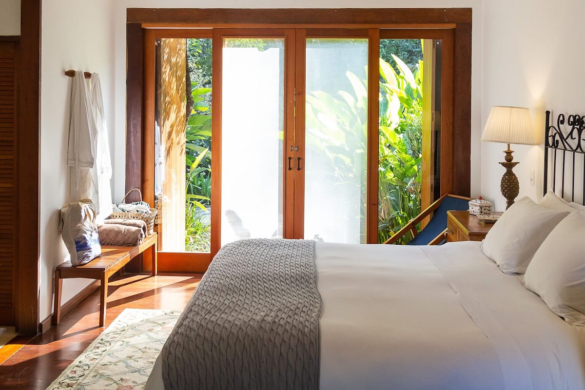 Une chambre avec un grand lit, une table de chevet, une lampe, une fenêtre donnant sur un espace verdoyant et un appareil de climatisation.