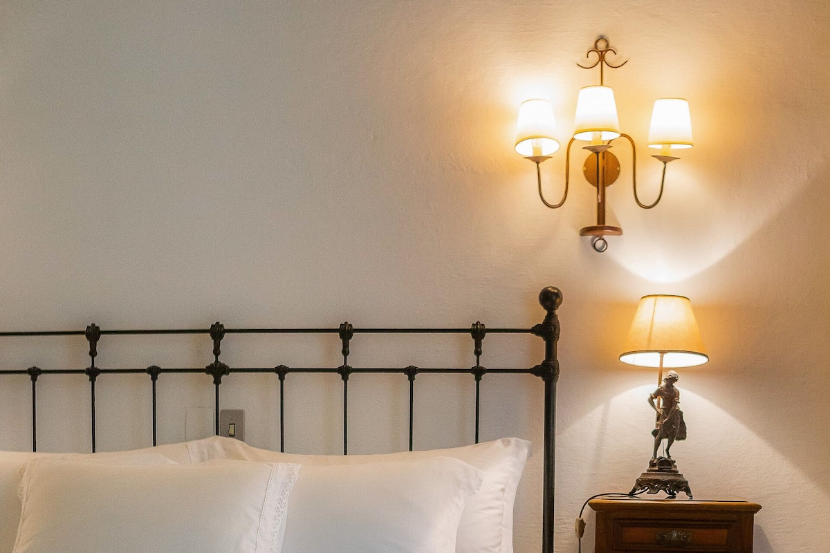 Un lit bien fait, avec du linge de lit blanc, une petite table de chevet surmontée d’une lampe, et un tapis à motifs sur le plancher en bois.