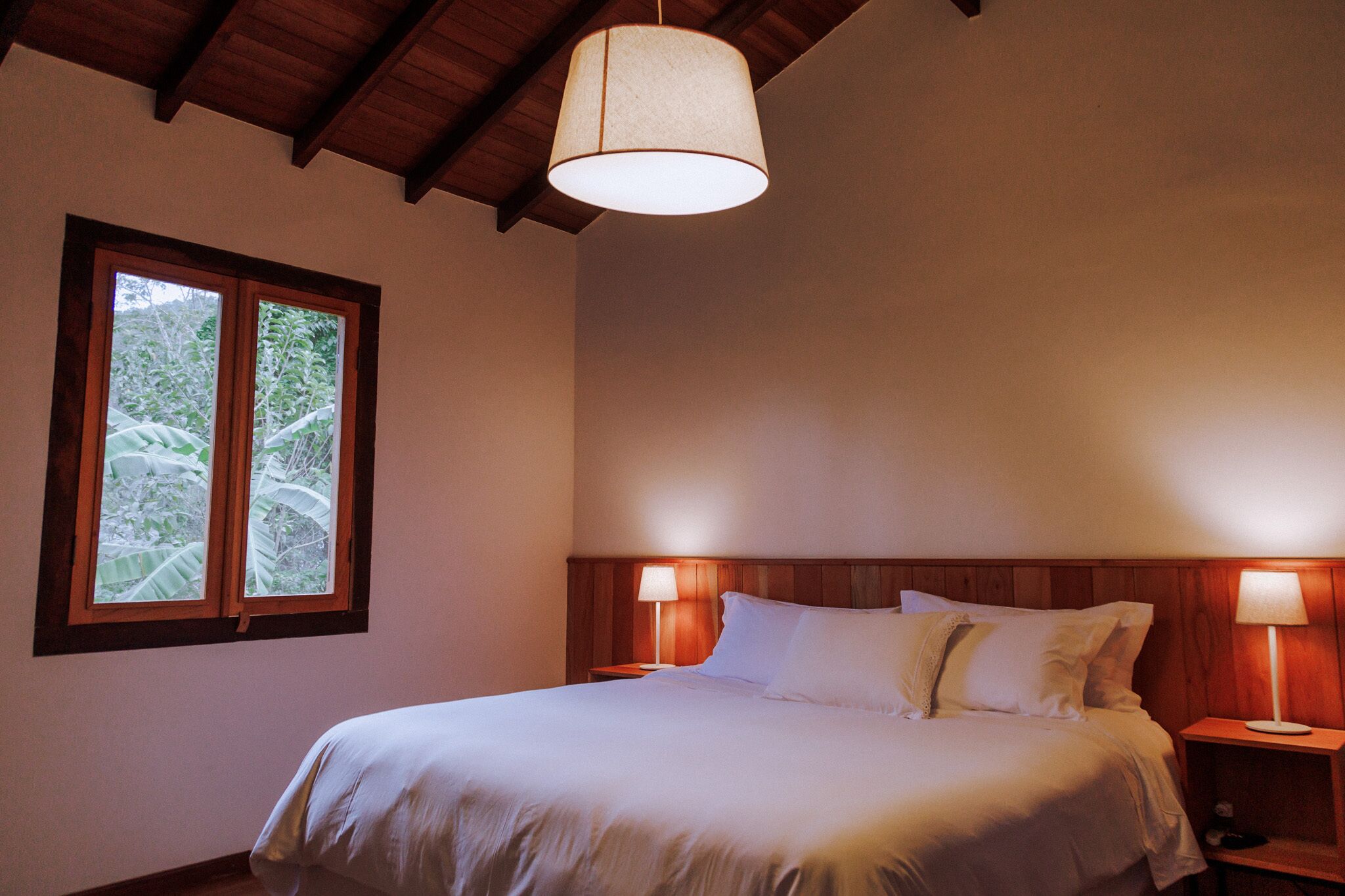 A bedroom with a bed, two bedside lamps, a window with a view of greenery, and a wooden ceiling.