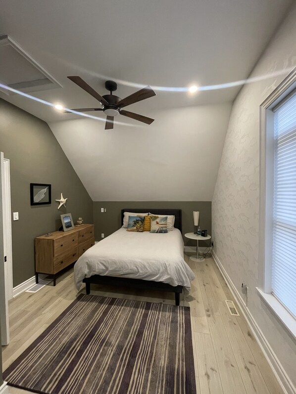 Une chambre avec un plafond en pente, un lit avec de nombreux oreillers, une commode en bois, une petite table de chevet avec une lampe et une grande fenêtre avec des stores.