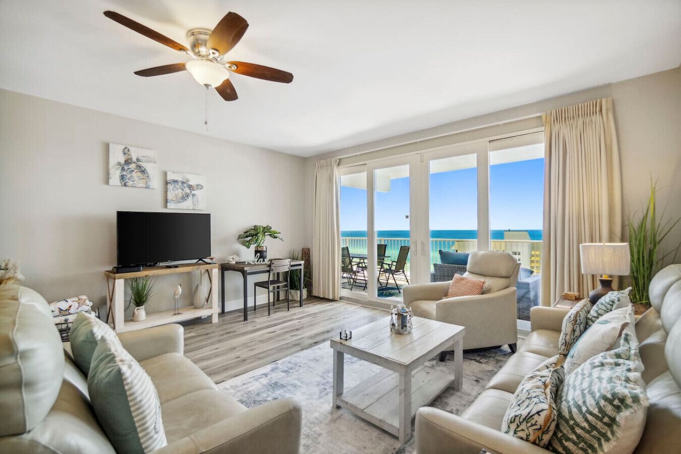 A spacious living room with a flat-screen TV, comfortable seating, a ceiling fan, and a large window overlooking the ocean.