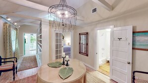 Un couloir bien éclairé, agrémenté d’un lustre, avec une table ronde décorée de bougies, et une vue sur une chambre à coucher où l’on aperçoit un lit et une fenêtre.