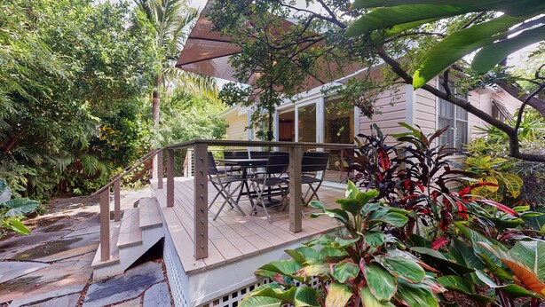 Une terrasse en bois, agréée de mobilier d’extérieur, entourée d’une végétation luxuriante, avec une maison en arrière-plan.