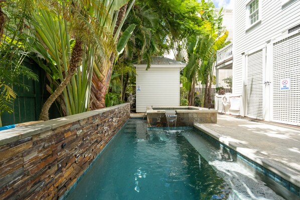 A swimming pool with a stone wall, surrounded by lush greenery and a small white building.