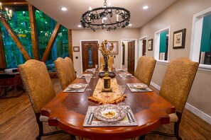 A dining room with a large wooden table set for six, surrounded by upholstered chairs with floral patterns. A chandelier hangs from the ceiling, and a large window offers a view of trees. - This Gives a “Cabin” a Whole New Meaning! (Carbondale)