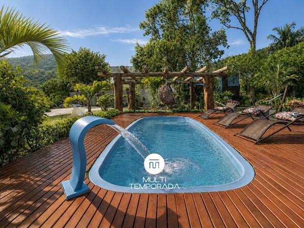 A swimming pool with a wooden deck, surrounded by lush greenery and a pergola structure. - Casa com Piscina em Gov Celso Ramos - SC (Governador Celso Ramos)
