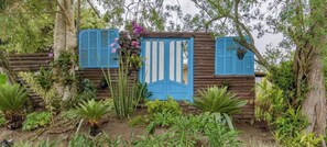 A rustic wooden structure with a blue door and shutters, surrounded by lush greenery and tropical plants. - Casa com Piscina em Gov Celso Ramos - SC (Governador Celso Ramos)