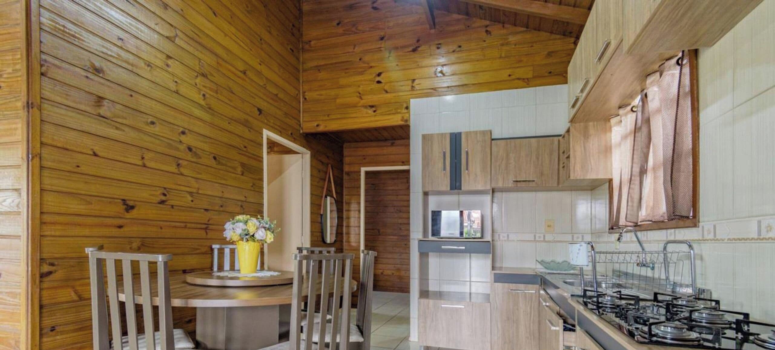 A wooden interior with a kitchen area, including a dining table with chairs, a small round table with a vase, and a stove.