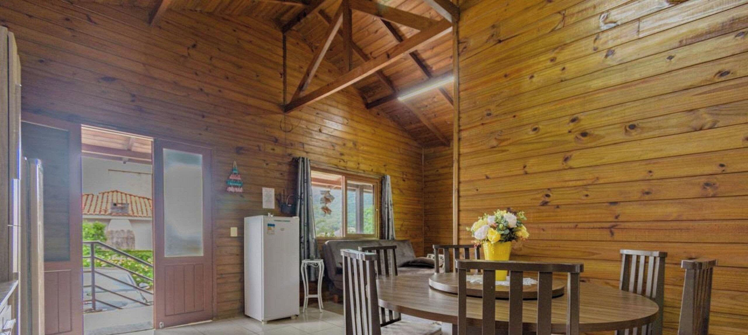 Interior de cabana de madeira com mesa de jantar, cadeiras, geladeira e janela.