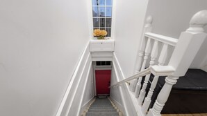 A staircase with a red door at the bottom, white balusters, and a small window with a view of buildings. - The Boat House Loft | Waterfront Rooftop Terrace (Belleville)