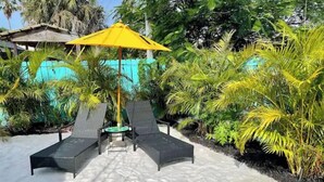 A sunlit outdoor seating area with two lounge chairs and a small table under a yellow umbrella, surrounded by lush tropical plants and a turquoise wall.