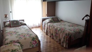 A room with two single beds, each with a floral bedspread and a decorative pillow. There is a window with sheer curtains, a wooden cabinet above the beds, and a tiled floor. - Apartment on the 2nd Floor 5 min from the spa (Saint-Vincent)