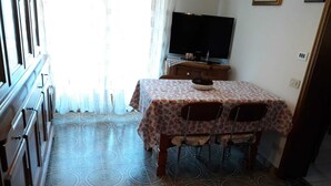 A dining area with a table covered by a floral tablecloth, two chairs, a television on a stand, and patterned floor tiles. - Apartment on the 2nd Floor 5 min from the spa (Saint-Vincent)