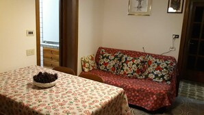 A dining area with a table covered in a floral tablecloth, a sofa with a floral cushion, and a wall with framed pictures. - Apartment on the 2nd Floor 5 min from the spa (Saint-Vincent)