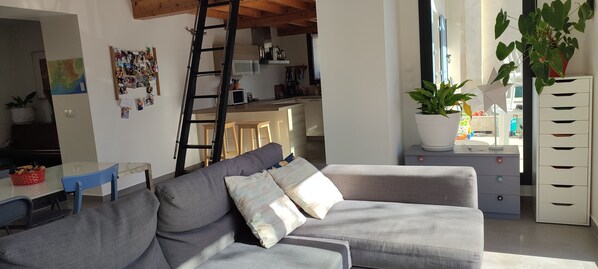A living room with a grey sofa, a small table with a basket of fruit, a ladder, a wall with photos, and a kitchen in the background. - Modern house on a large, wooded lot in a quiet location (Gréasque)