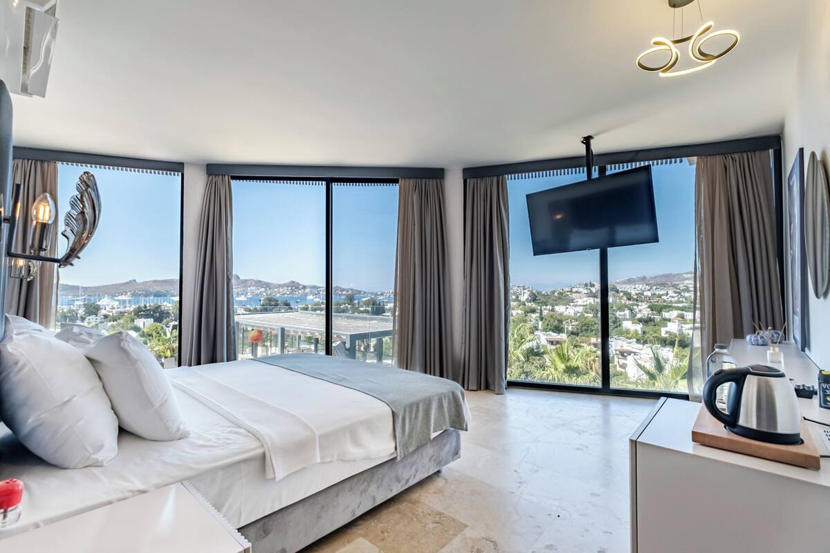 A modern hotel room with a large bed, a flat-screen TV, and a view of a cityscape through large windows.