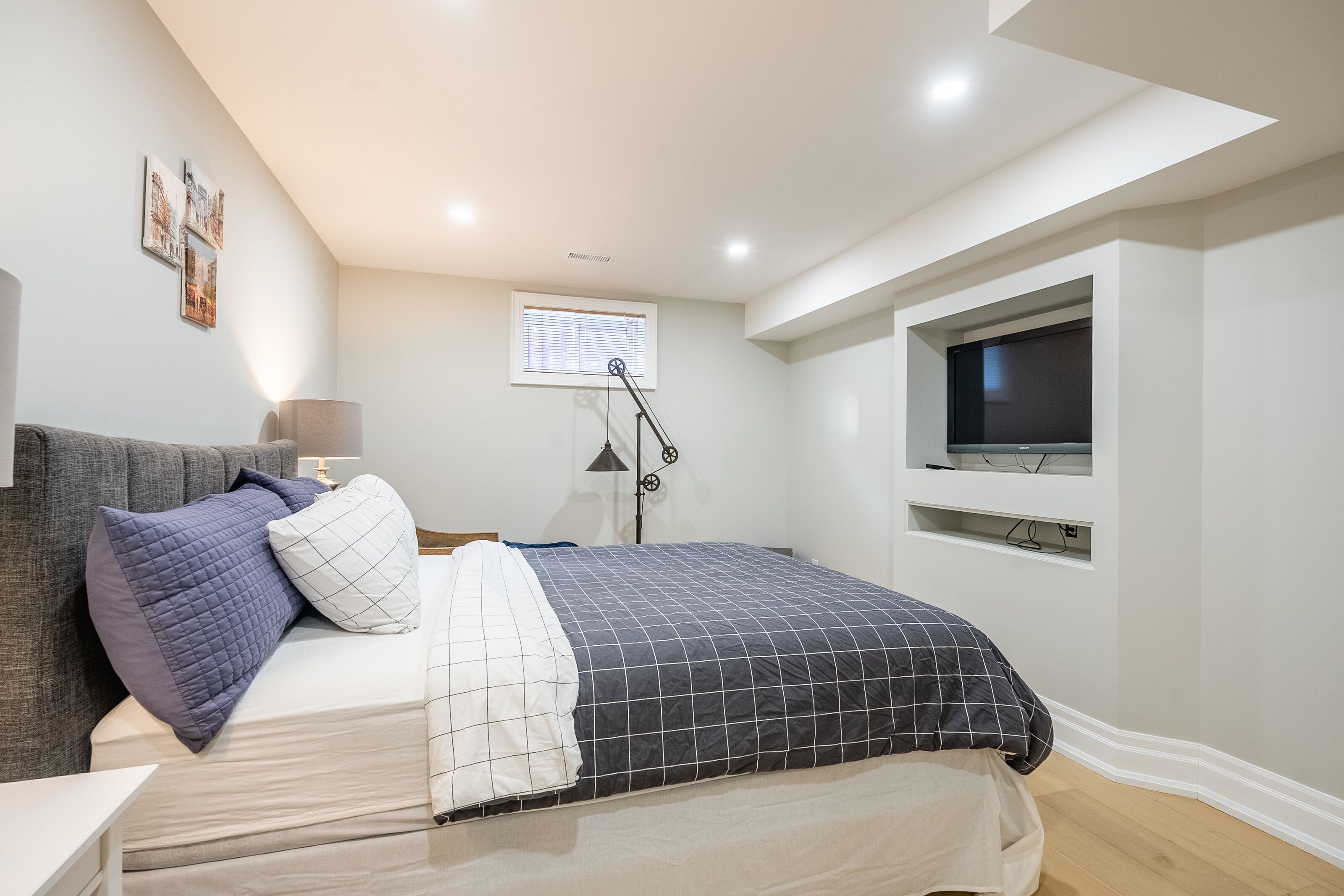 Une chambre à coucher moderne avec un lit, un téléviseur fixé au mur, une fenêtre avec des stores et une lampe.
