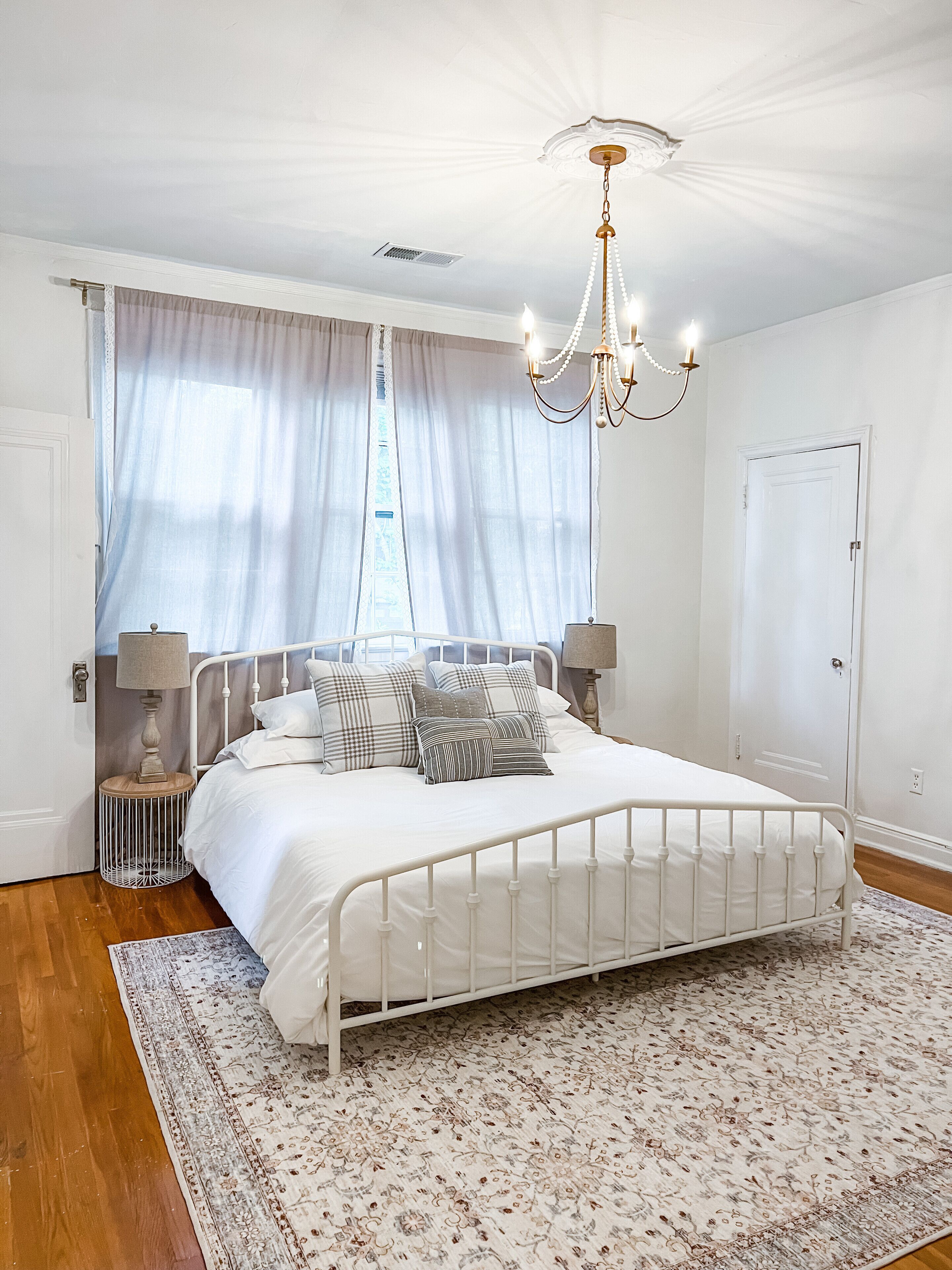 A bedroom with a white metal bed frame, a patterned rug, a chandelier, and a window with sheer curtains.