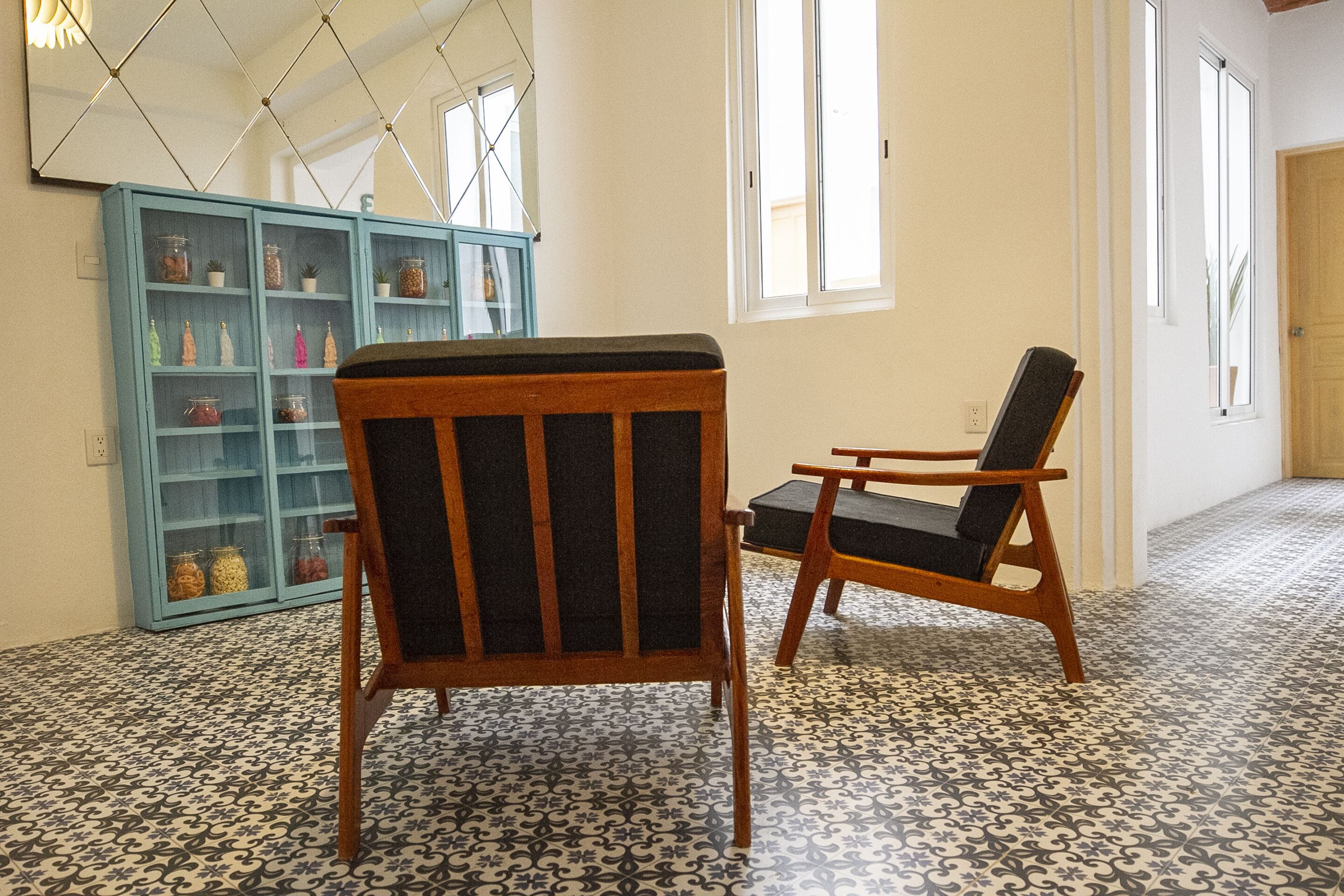 A room with patterned floor tiles, a wooden chair with a cushion, and a blue cabinet with glass doors containing jars and small plants.
