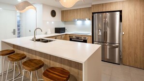 A modern kitchen with a large island, stainless steel appliances, wooden bar stools, and pendant lighting. - Unit 4 Sundeck Gardens (Sunshine Coast)