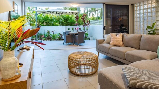 A modern living room with a sofa, a round wicker ottoman, and a dining area with a table and chairs. - Unit 4 Sundeck Gardens (Sunshine Coast)